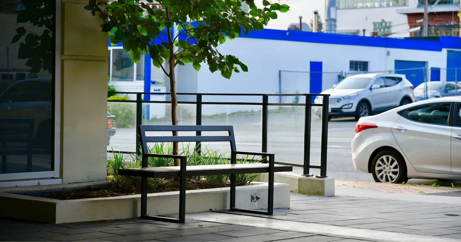 A Skyline bench provides seating in a commercial area with a planter box. The bench features a black frame and light-colored recycled plastic lumber.