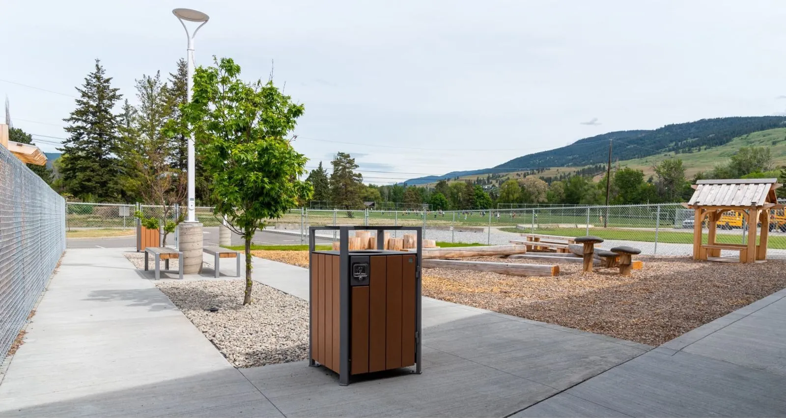 Skyline waste receptacle is placed near benches at the YMCA in Lake Country, British Columbia. The setting features a playground and a view of the surrounding hills.