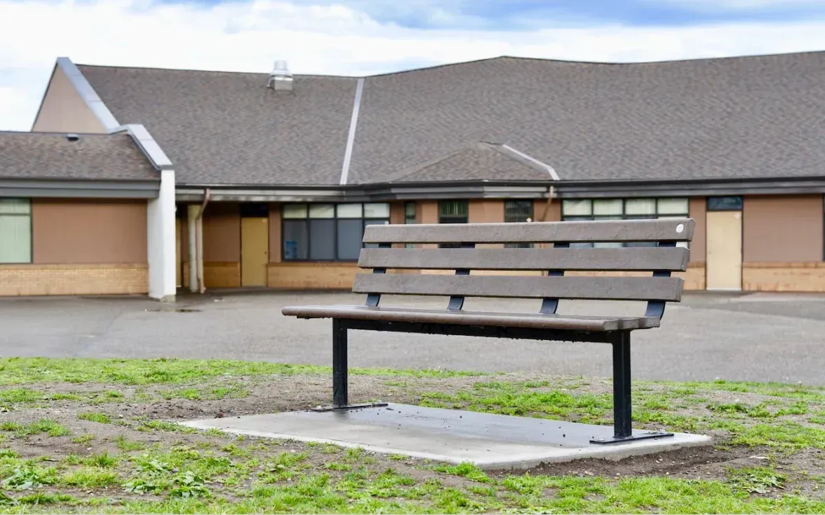 A Standard Bench provides seating outside a school building. The bench features recycled plastic lumber slats and a powder-coated metal frame.