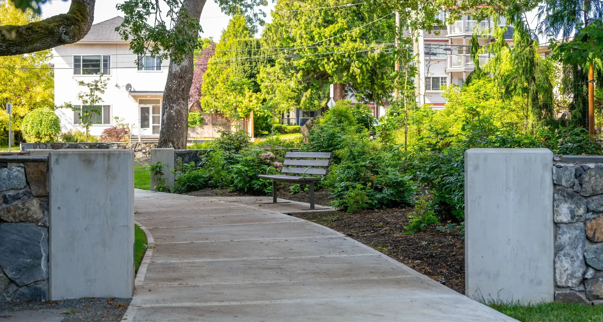 A Standard Bench with armrests sits along a concrete path in a landscaped park setting. The bench features recycled plastic lumber.