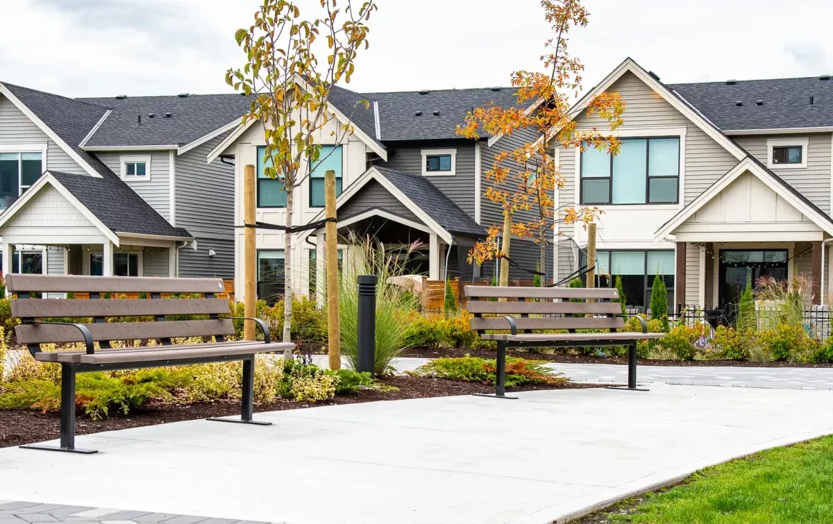 Standard benches provide seating in a landscaped area with modern townhouses. The benches feature recycled plastic lumber and powder-coated metal frames.