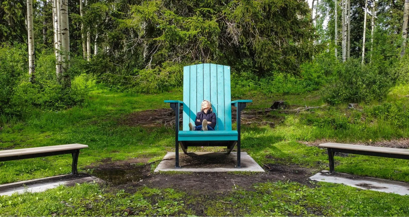 Oversized Adirondack chair in a park-like setting. The chair is painted in a bright blue color and is surrounded by lush greenery and trees.
