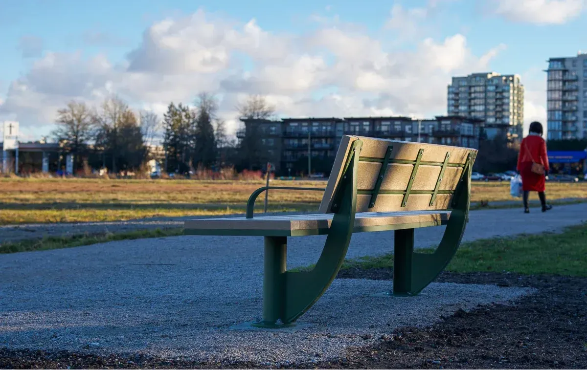A SurRe bench offers seating along a gravel path in a park setting. The bench features recycled plastic lumber and a powder-coated frame.