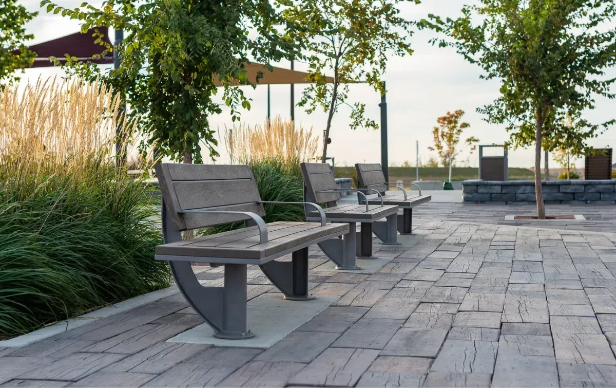 Three SurRe benches line a paved walkway in a landscaped park setting. These benches offer comfortable seating with a modern design.