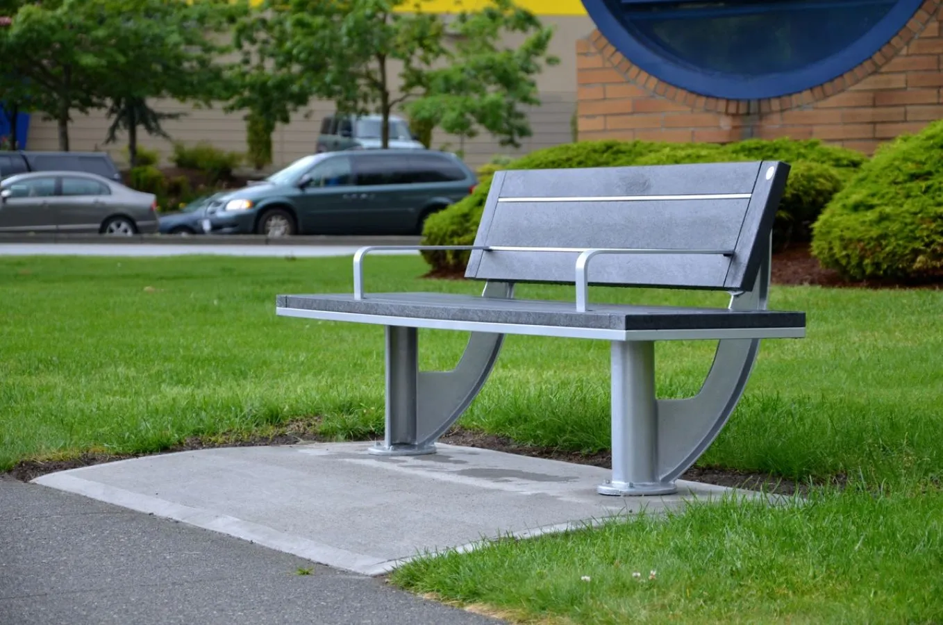 A SurRe park bench provides seating in a grassy area next to a sidewalk. The bench features recycled plastic lumber and a powder-coated metal frame.