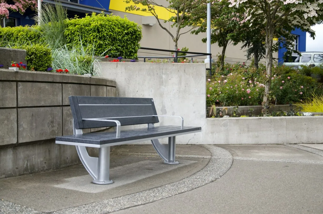 A SurRe bench provides seating in a landscaped area with concrete walls and colorful building in the background. The bench features recycled plastic lumber and a powder-coated metal frame.