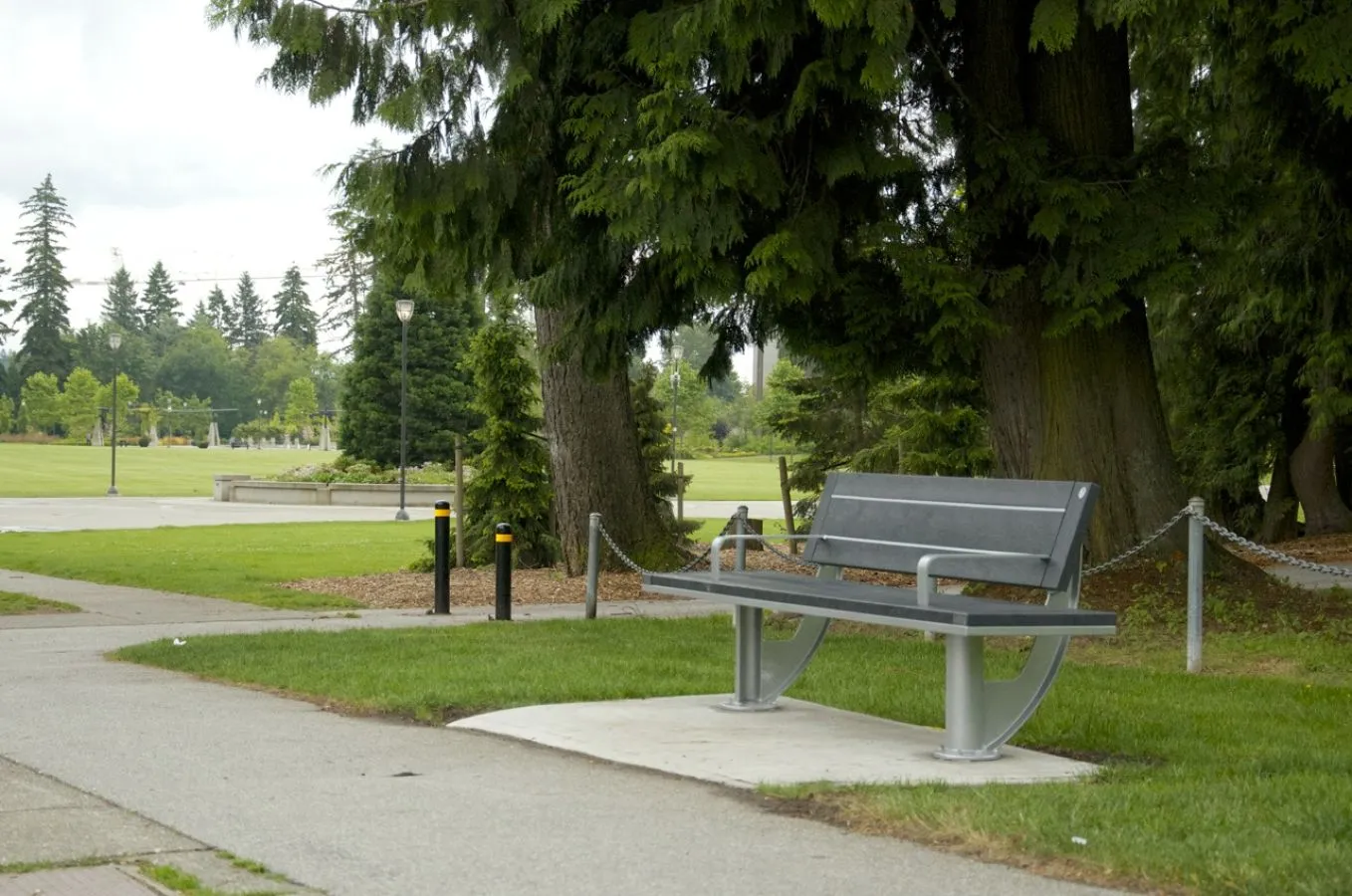 A SurRe bench sits on a concrete pad next to a paved path in a park setting. The bench features recycled plastic lumber and a metal frame.