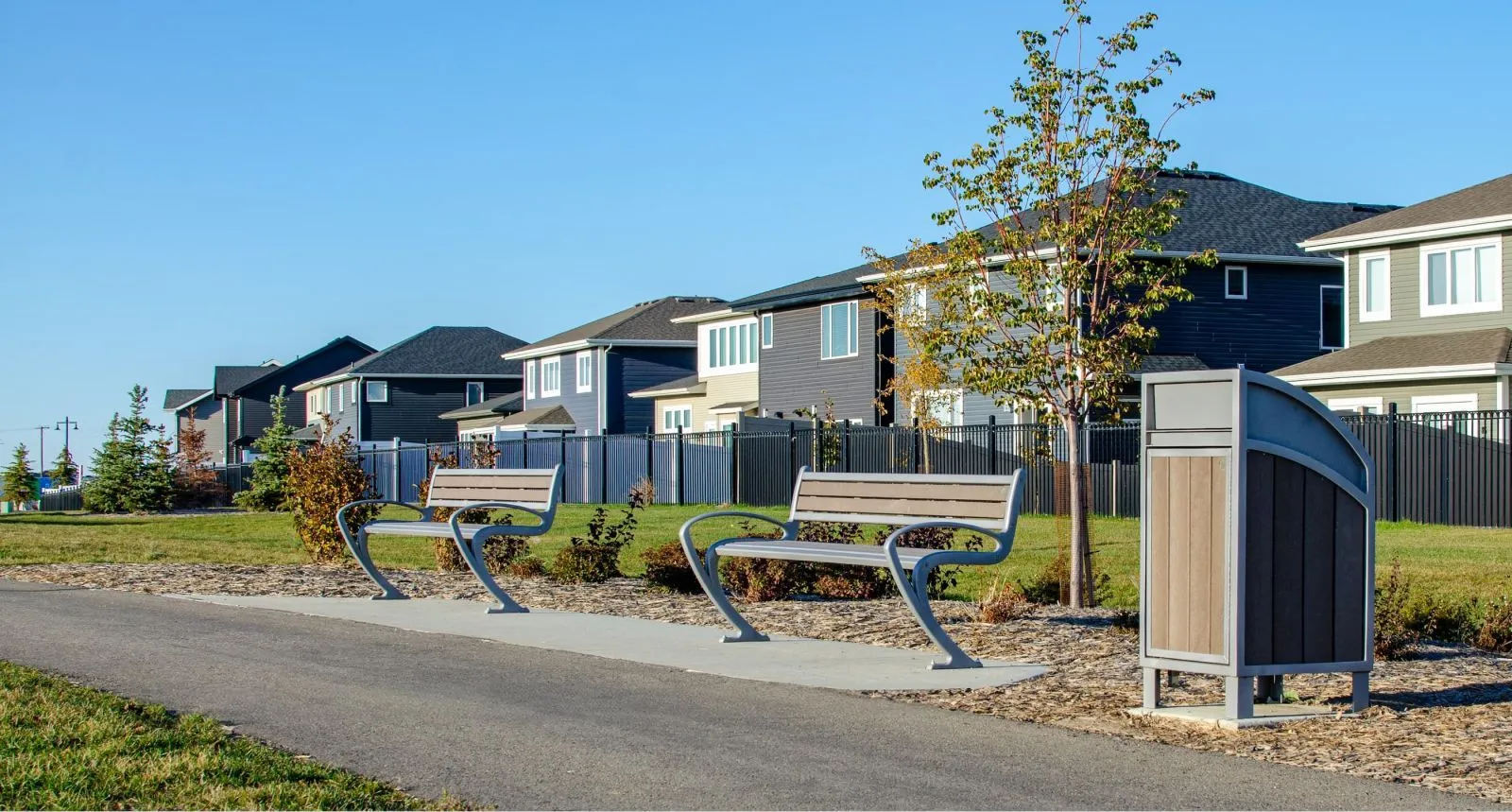 TENAJ benches and a Modena Curved Top waste receptacle provide seating and waste disposal along a paved path in a residential community. The site furniture complements the modern architecture and landscaping of the Brighton neighborhood.
