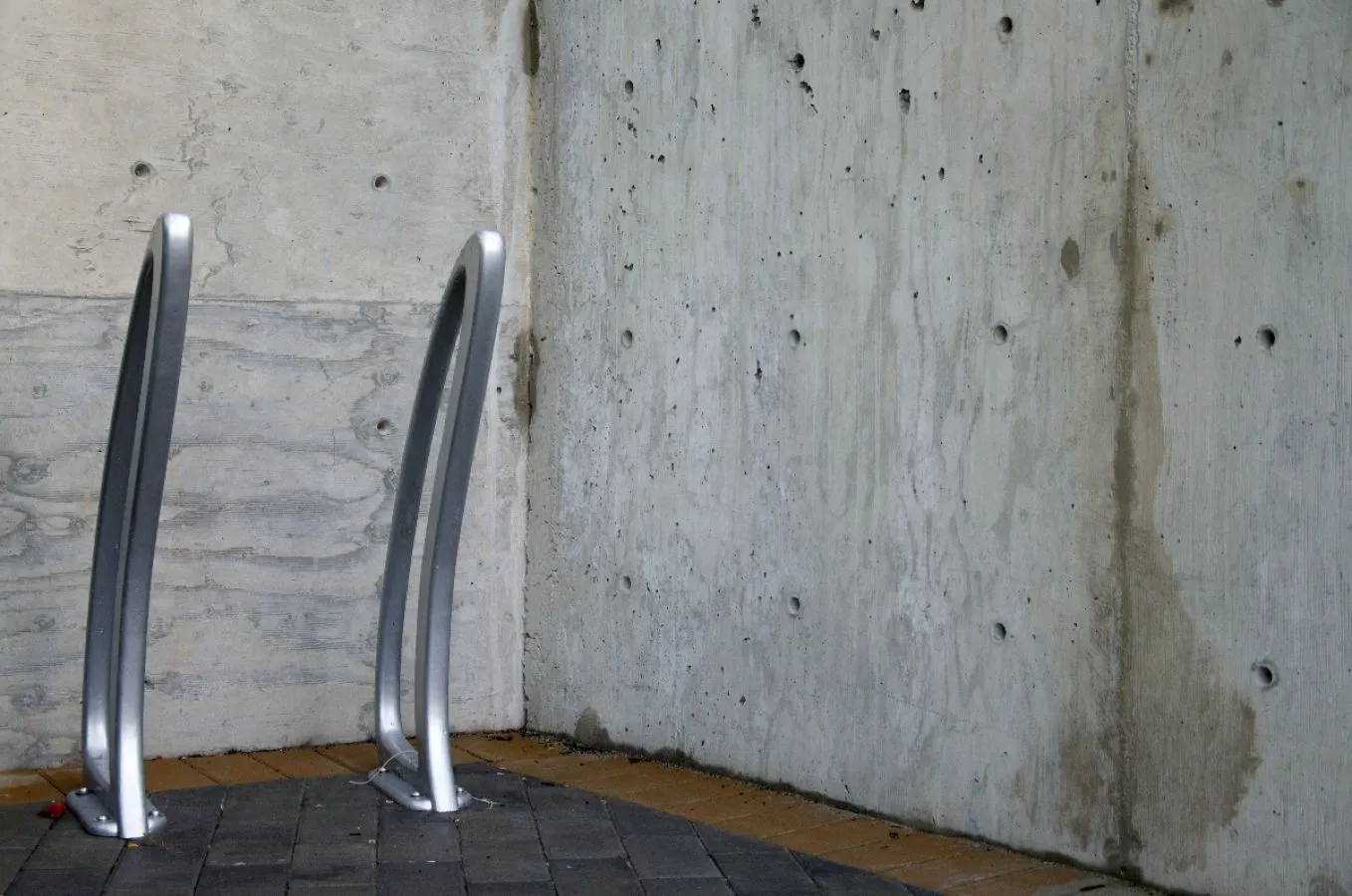 Two TENAJ bike racks stand against a concrete wall. These durable racks provide secure bike parking for commercial or public spaces.