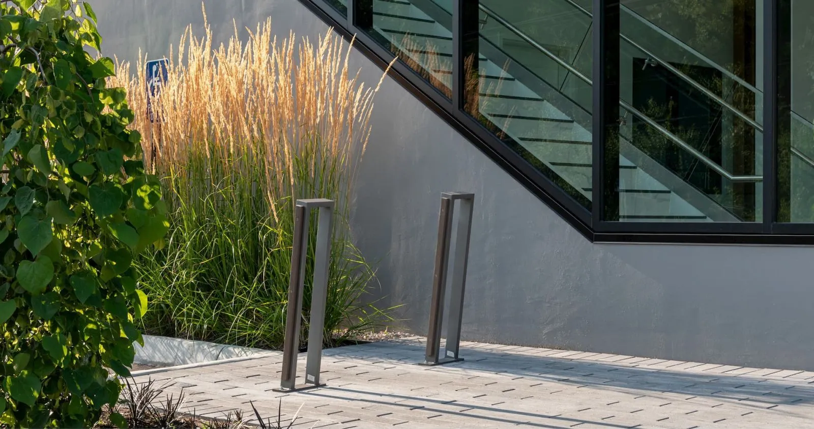 Two Urban Form 2 Space Bike Racks stand on a paved area next to a modern building and ornamental grasses. These bike racks provide convenient and secure parking for bicycles.