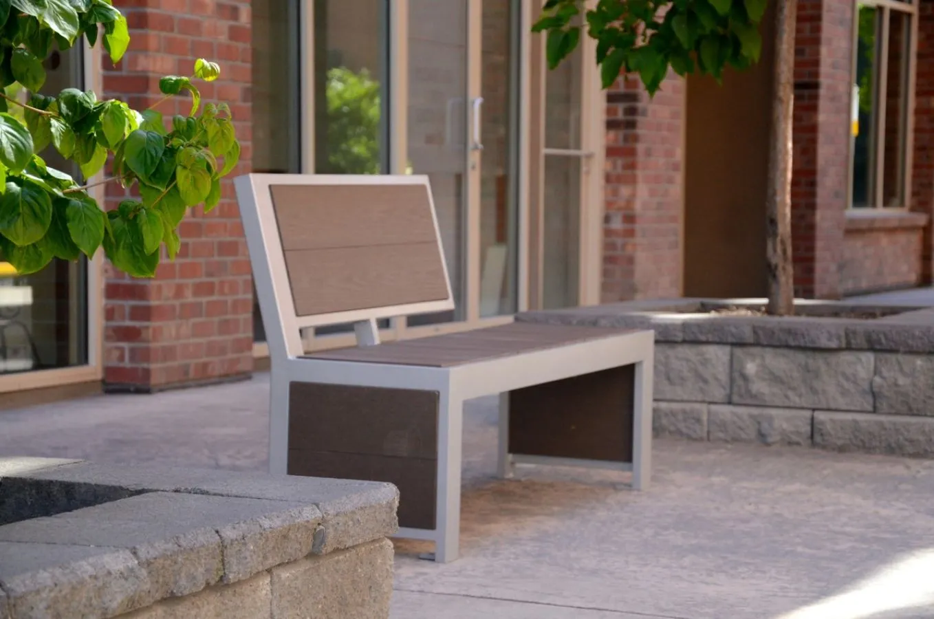 Urban Form bench with a powder-coated metal frame and recycled plastic lumber seat and back. The bench is situated on a paved area with a brick building in the background.