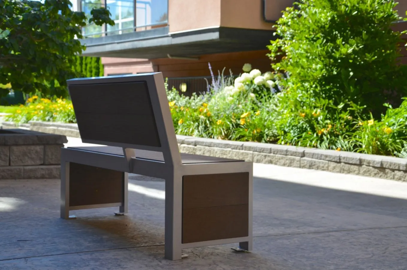 Urban Form bench with a backrest, featuring recycled plastic lumber and a powder-coated metal frame, sits in a landscaped area. This modern bench provides comfortable seating in an outdoor setting.