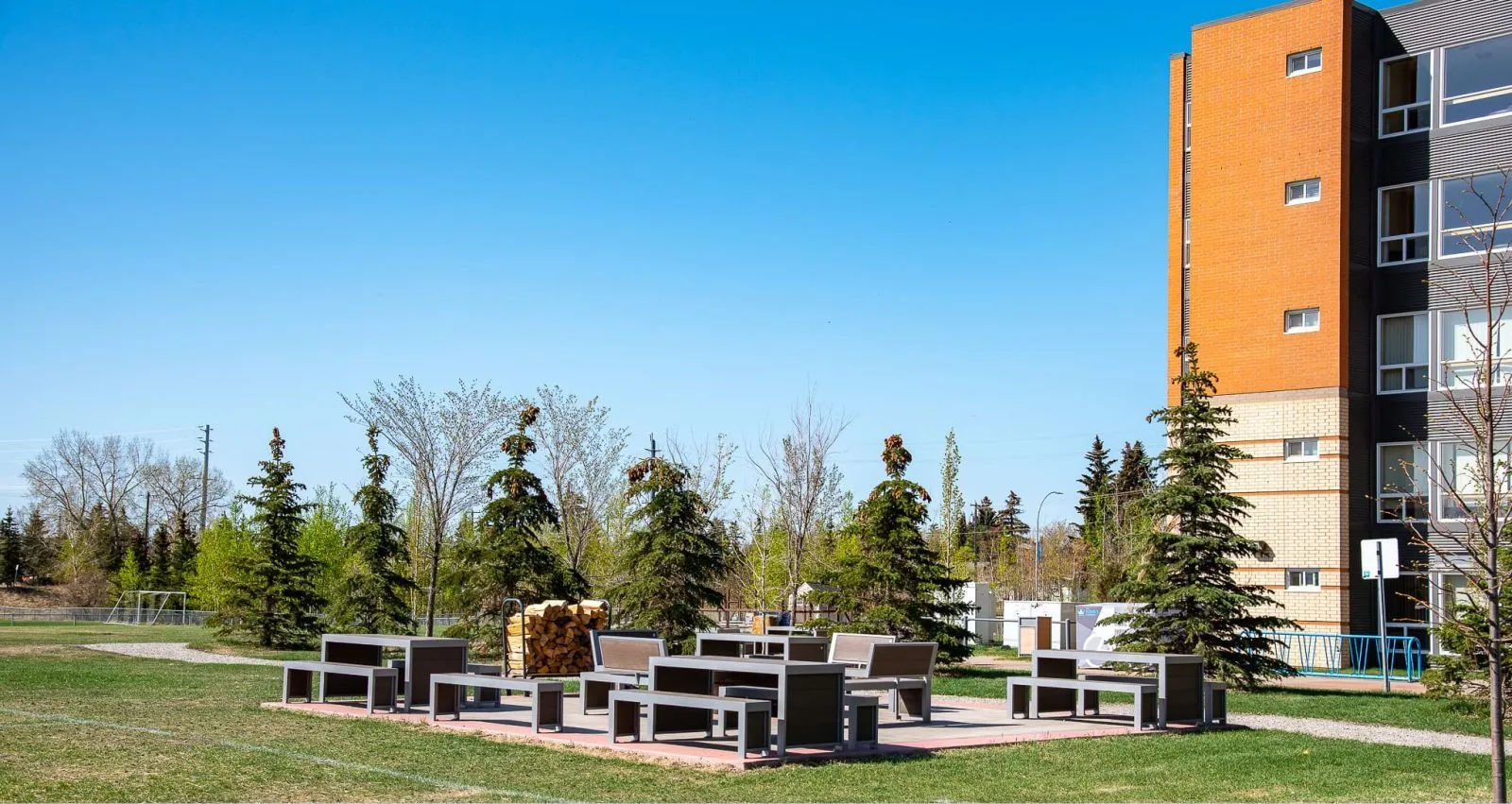 Outdoor picnic area with Urban Form picnic tables and benches on a paved pad near a university building. The site furniture features recycled plastic lumber and powder-coated metal frames.