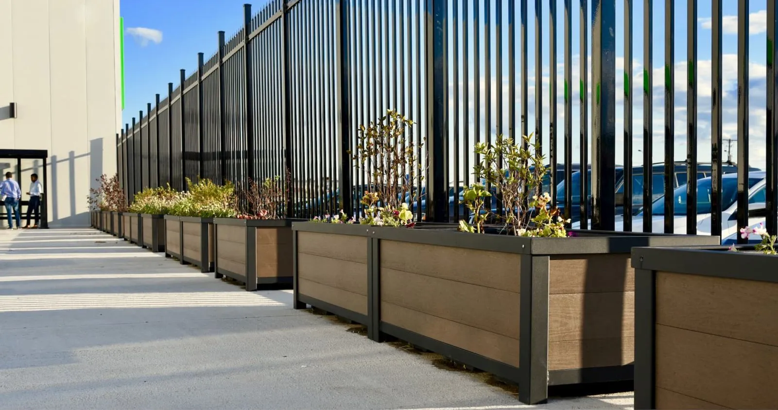 A row of self-watering rectangular planters line a walkway, adding greenery to the space. The planters feature recycled plastic lumber and a dark powder-coated metal frame.