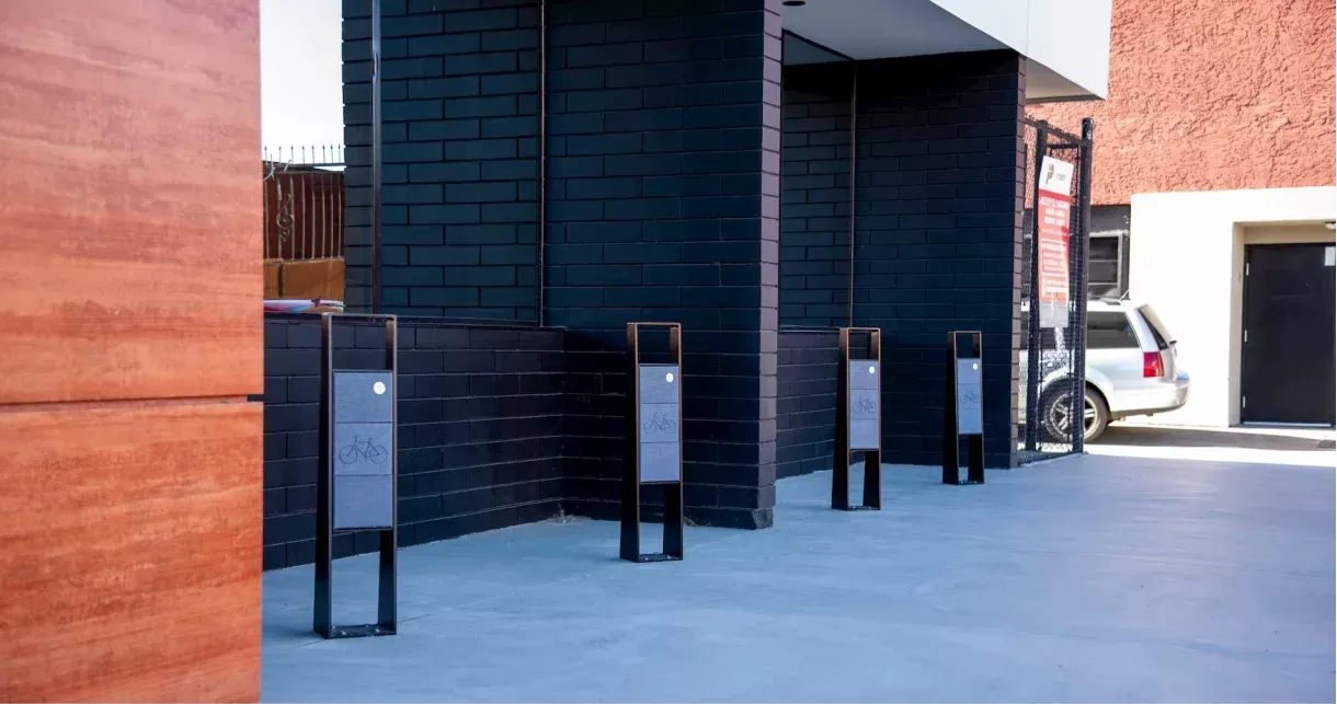 Bayview 2 Space Bike Racks provide secure bicycle parking outside a commercial building. The bike racks feature a powder-coated metal frame and a panel with a bicycle graphic.