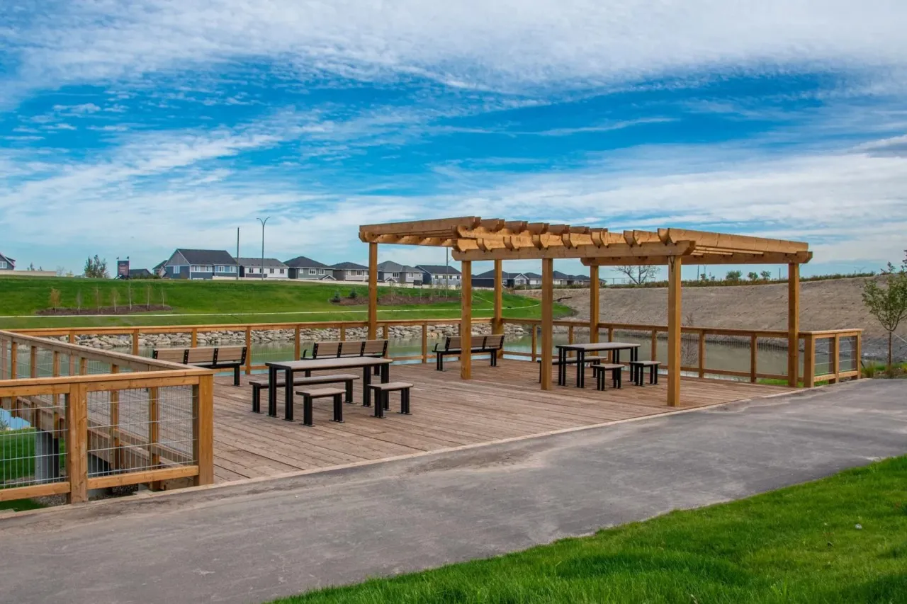 Outdoor picnic tables and benches provide a gathering space on a wooden deck overlooking a pond. The setting includes a pergola for shade and a walking path.