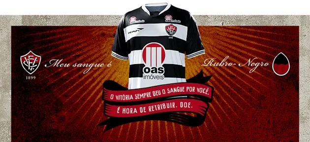 A imagem mostra uma camiseta do uniforme do Esporte Clube Vitória. A camiseta tem listras horizontais, intercalando entre as cores preto e branco. Na imagem, pode-se ler o texto: "Meu sangue é rubro-negro. O Vitória sempre deu o sangue por você. É hora de retribuir. Doe".