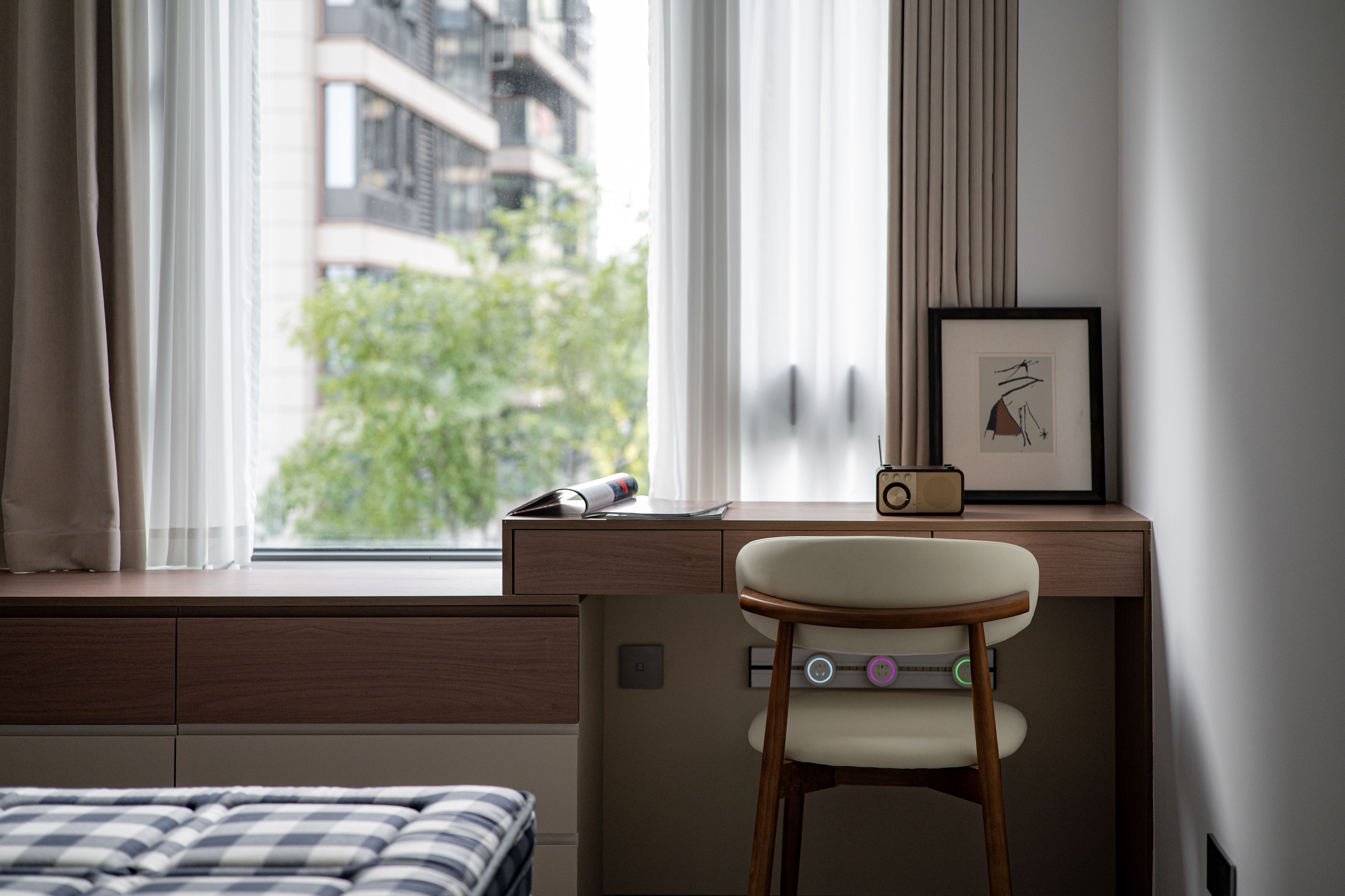 A bright home office with a desk positioned near a large window, allowing natural light to illuminate the workspace. The room features light curtains 