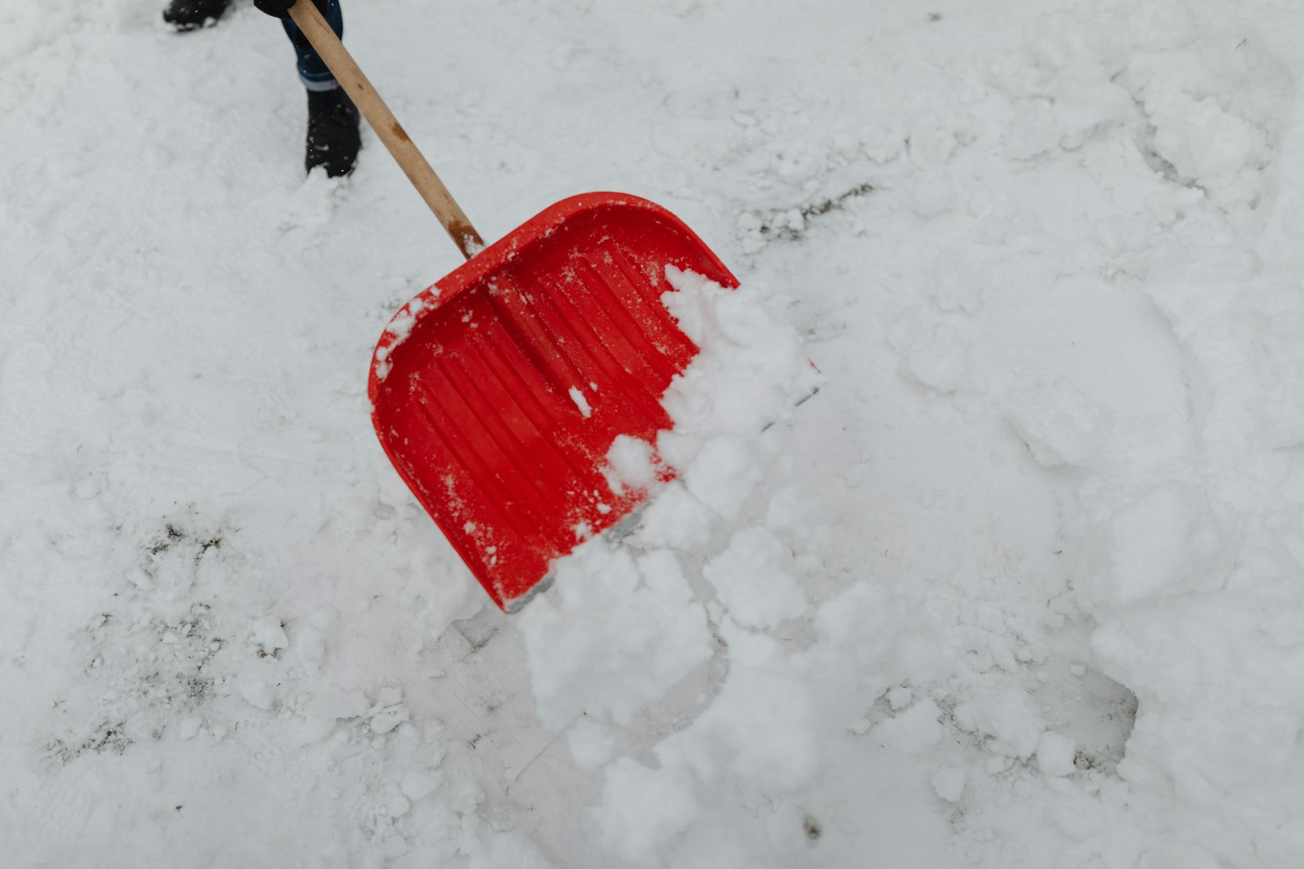 A person using a shovel to clear snow from a walkway, with a snow blower in the background, ensuring property safety during winter.
