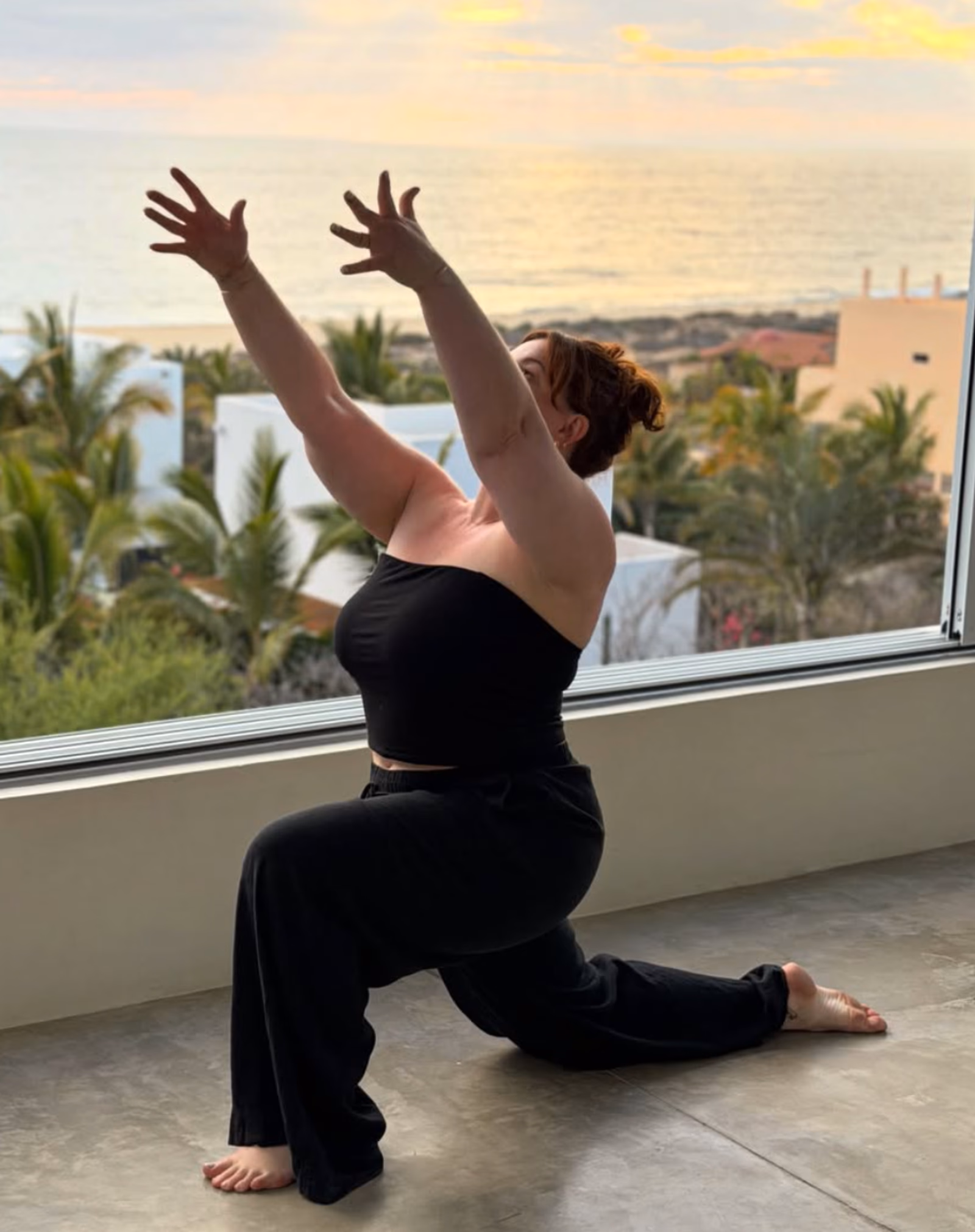 Elise in a low lunge yoga pose by a window overlooking the coast