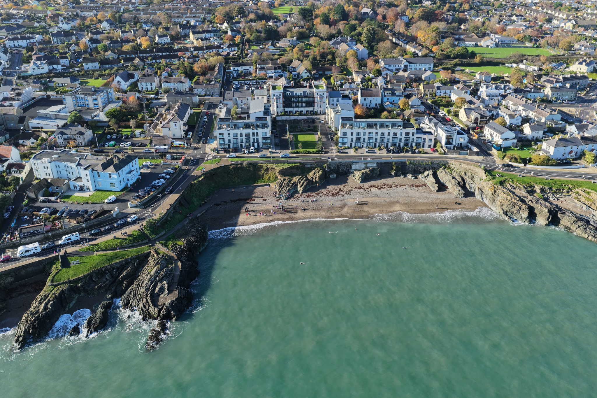 An aerial drone perspective of Greystones' coastal cove, featuring swimmers in clear turquoise water, a sandy pebble beach, and a cliffside promenade.